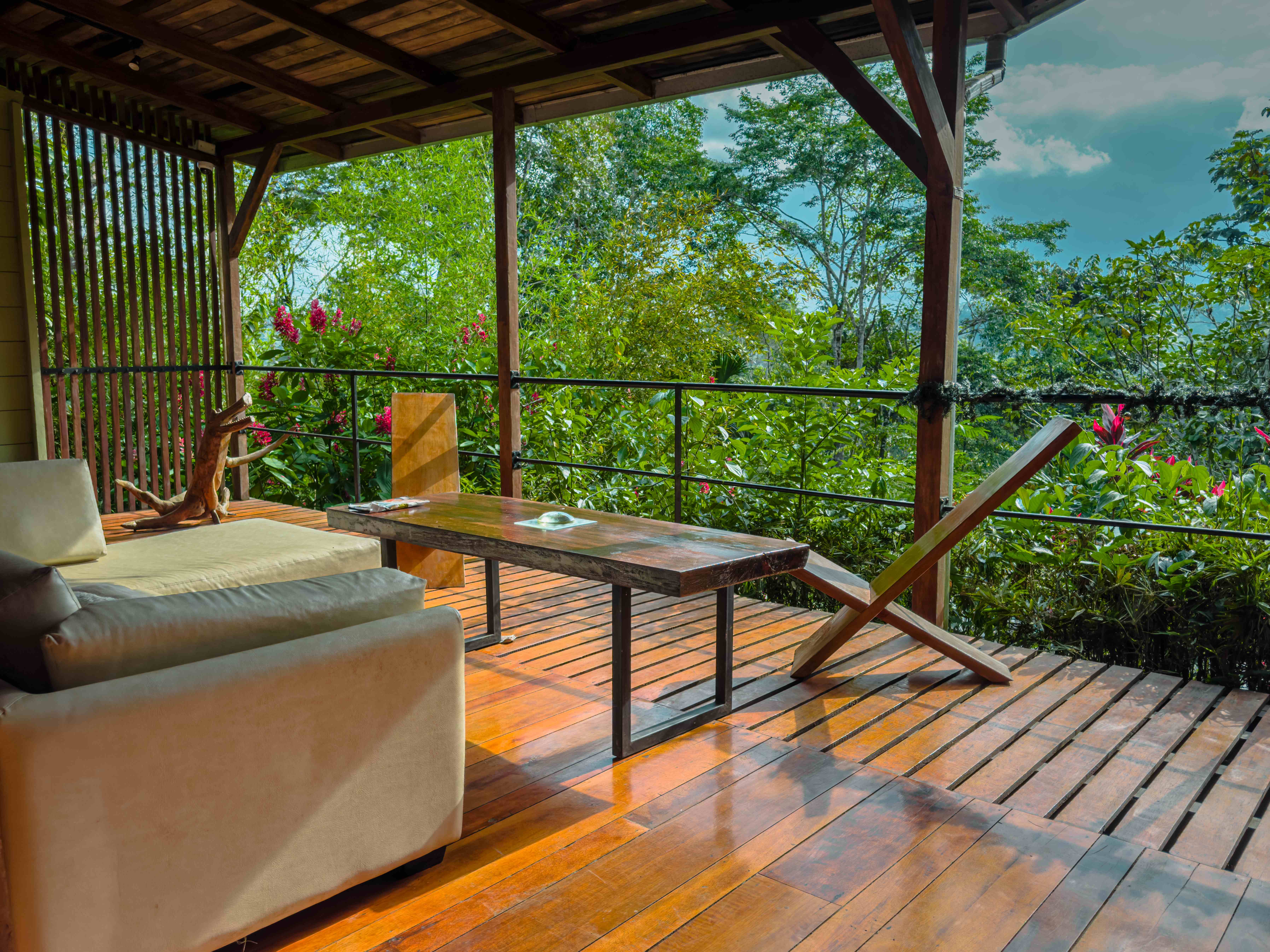 Wooden terrace lounge of a jungle lodge with a sofa, coffee table, and folding chair overlooking dense rainforest