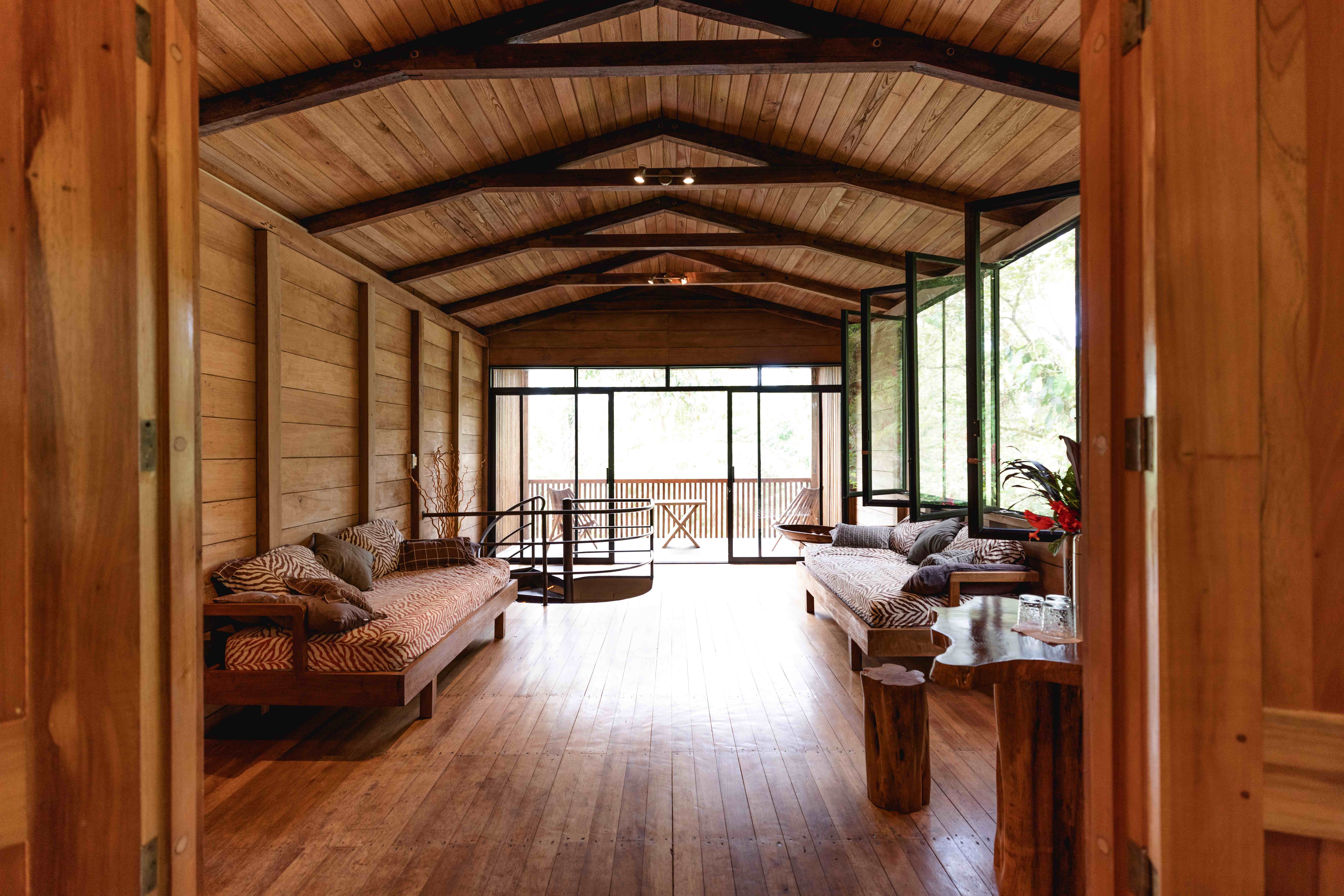 Open-air timber lodge great room with vaulted beamed ceiling, zebra-print daybeds, and a spiral staircase leading to a jungle-view balcony